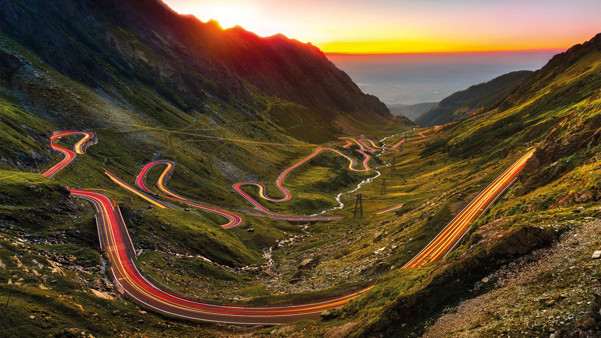 Traffic light trails on windy mountain roads leading towards the sea and sunset