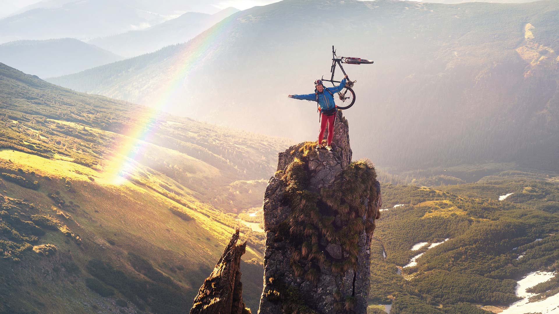 Person on top of a high mountain rock and holding their bike up, with a lovely rainbow in the background