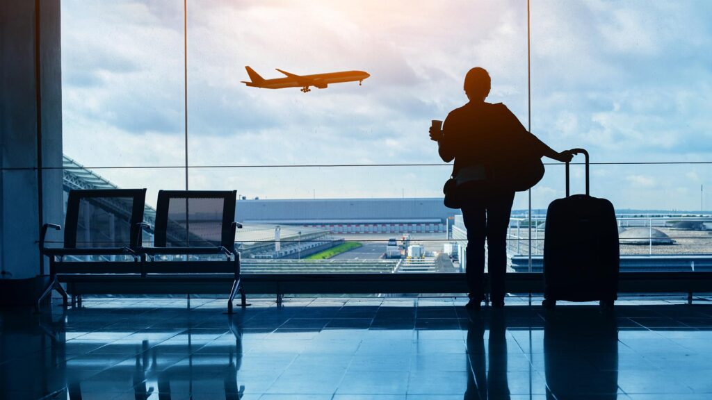 Person waiting at airport with their suitcase, looking through window at plane taking off